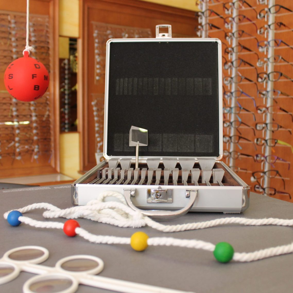 Maletín con prismas ópticos, junto a una cuerda con bolas de colores, una bola roja con letras negras y un flipper acomodativo.
