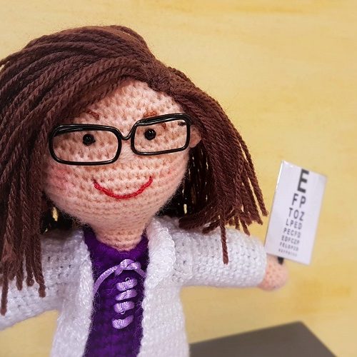 Muñeca de lana con cabello castaño, gafas y un optotipo en la mano representando una optometrista.